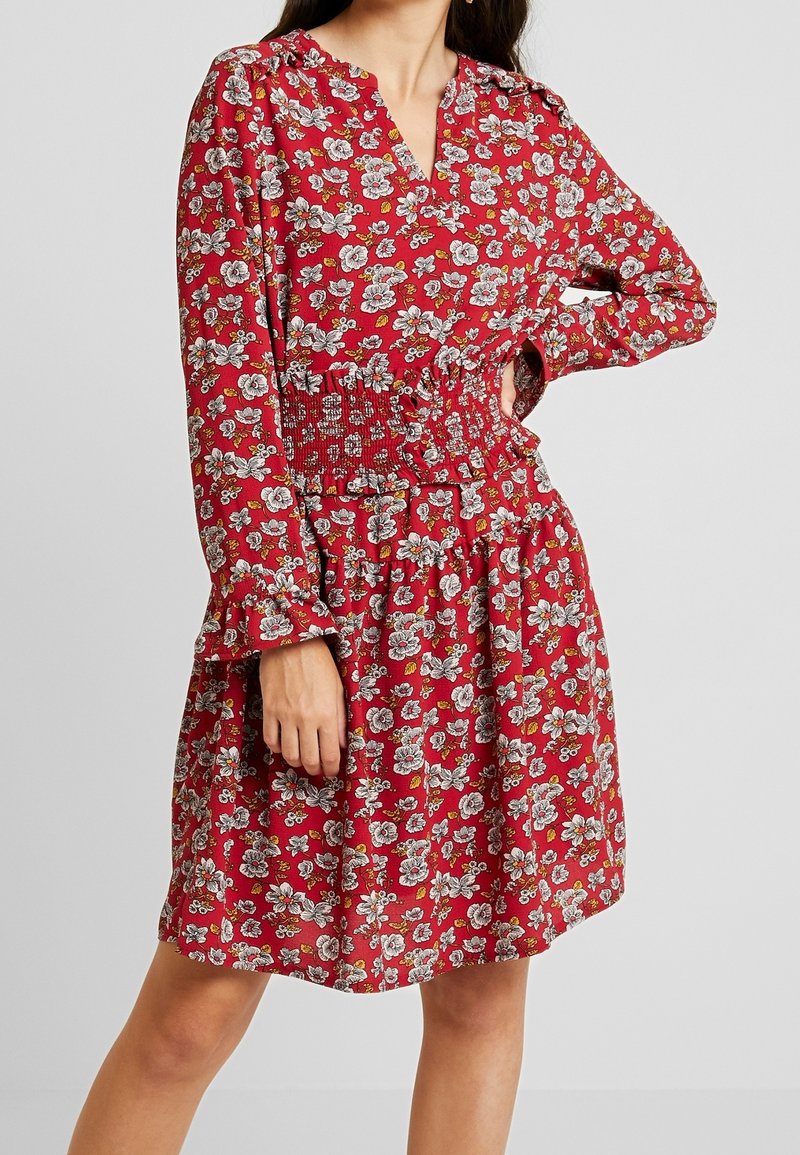 Femme portant une robe rouge à fleurs à manches longues avec une taille smockée et un décolleté en V, debout, une main sur la hanche, devant un fond uni.