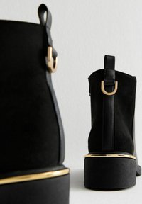 Botas negras hasta el tobillo con un acabado de ante, que presentan detalles en cuero liso y un detalle metálico dorado cerca de la suela. Diseño de tacón grueso.