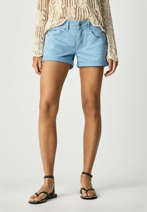 Kvinde iført lyseblå shorts med opkantsøm, beige strikket langærmet top og sorte ankelremssandaler med nitter, stående mod en ensfarvet baggrund.