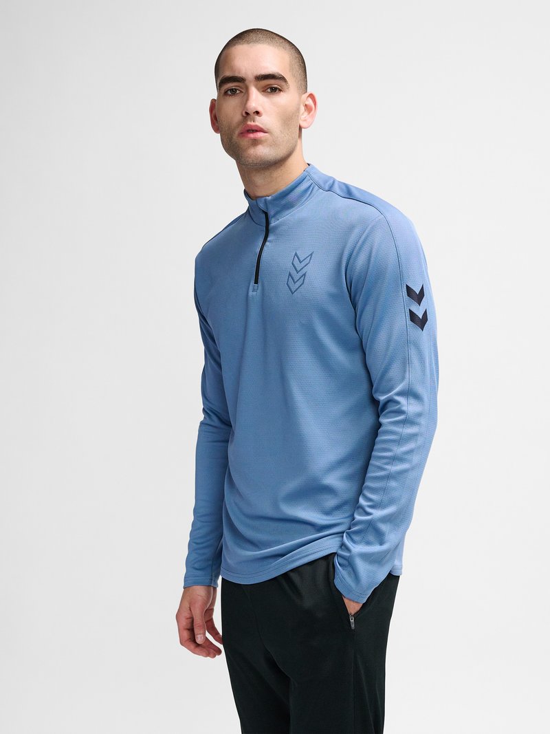Jonge man die een blauwe, lange mouwen sporttrui met een kwart rits draagt met zwarte chevron-logo’s op borst en mouw, zwarte broek, staand tegen een witte achtergrond.