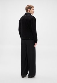 Melnas zamša jaka ar standarta apkakli, aizmugures yoke un pogu aizdari; kombinēta ar platām melnām biksēm, skatīta no aizmugures.