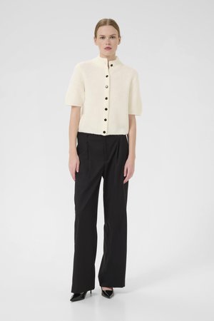 Cream short-sleeve knitted cardigan with a high collar and black buttons, paired with black straight-leg trousers and pointed black heels.