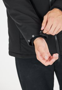Sort pufferjakke med lynlås og trykknapmanchetter. Nærbillede af hænder, der justerer manchetterne. Tekstureret stof og tætsiddende design.