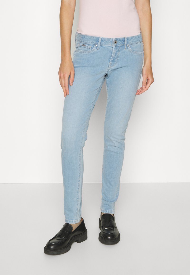 Jean skinny bleu clair en denim, avec fermeture à bouton sur le devant, passants de ceinture et design à cinq poches. Portés avec des mocassins noirs.
