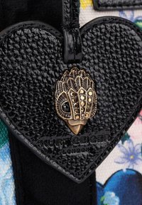 Etichetta a forma di cuore con texture nera, con un emblema decorativo tono oro con perline, fissata a una borsa di stoffa floreale e colorata.