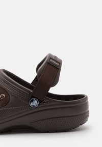 Sabot en caoutchouc brun avec sangle ajustable, surface texturée, accents circulaires et motif en relief sur les côtés. Détail du logo près du talon.