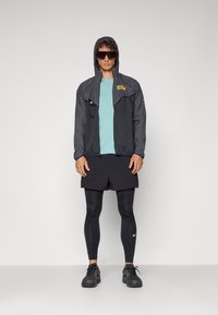 Athléta fekete szélálló dzsekit visel kapucnival, mentazöld pólót, fekete sortot és fekete leggingset fehér Nike logóval; napszemüveg is szerepel.