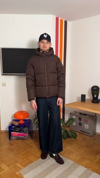 Dark brown puffer jacket with a hood, zip front, and padded design. Paired with wide-leg dark denim pants and dark shoes.