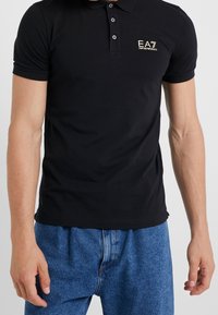 Svart pikétröja med korta ärmar och tvåknappslås. Har en liten EA7-logotyp i guld på vänster bröst. Tillverkad av bomull.