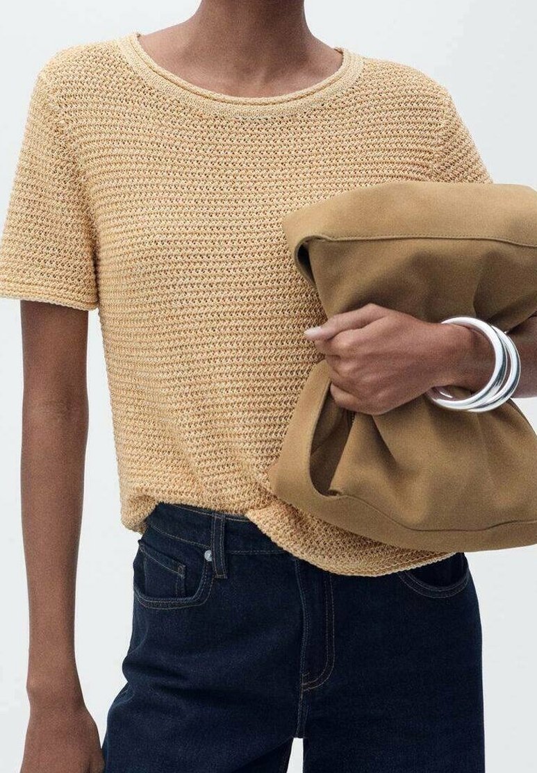 Personne portant un haut en maille beige à manches courtes et un jean foncé tient une grande pochette pliée couleur camel avec des bracelets argentés au poignet.
