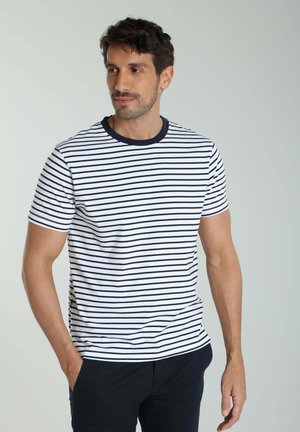Homme portant un t-shirt blanc avec des rayures noires horizontales et un pantalon noir, debout les mains dans les poches, regardant vers la gauche.