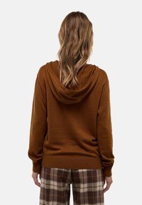 Sweat à capuche marron en tissu doux, avec un ourlet et des poignets côtelés. Porté avec un pantalon à carreaux, mettant en valeur une coupe décontractée et un style casual.
