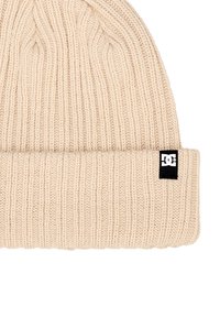 Pletená čapica v svetlej béžovej farbe s ryhovanou textúrou. Má prehnutý lem a malú čiernu nálepku s logom DC na boku.