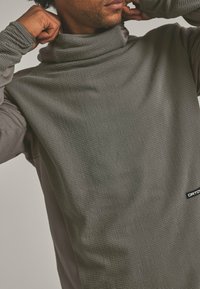 Mann, der den Kragen eines strukturierten grauen Langarmshirts mit einem kleinen schwarz-weißen Logo an der unteren Seite hochzieht.