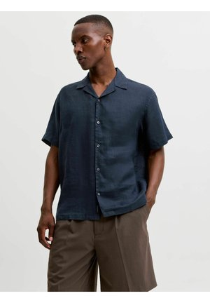 Man met kort haar die een donkerblauw, kort mouw, knoopjeshemd draagt en een bruine korte broek, staand met één hand in de zak tegen een effen achtergrond.