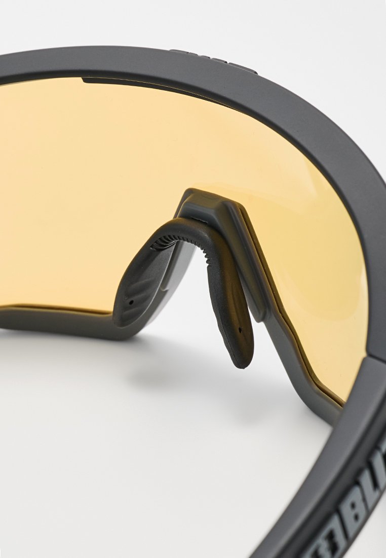 Nahaufnahme von schwarz gerahmten Schutzbrillen mit gelb getönten Gläsern und einem flexiblen Nasenpolster vor einem weißen Hintergrund.