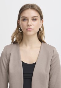 Jeune femme aux cheveux châtain clair, yeux bleus, portant des boucles d'oreilles argentées en créole, un haut noir et un blazer taupe, face à l'avant avec une expression neutre.