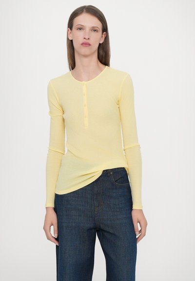Yellow long-sleeve henley top with a rounded neckline, button placket, and ribbed texture, paired with dark blue wide-leg jeans.
