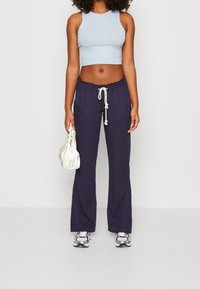 Un top court bleu clair, un pantalon en tissu navy avec cordon de serrage, et des chaussures de sport argentées. L'utilisateur tient un petit sac en satin crème avec cordon de serrage.