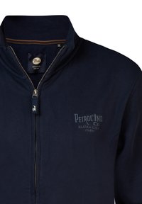 Marineblauwe zip-up jas met een hoge kraag, met een logo op de linkerkant van de borst en contrasterende stiksels op de kraag. De rits heeft een merklabel.