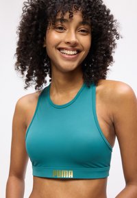 Χαμογελαστή γυναίκα με σγουρά μαλλιά που φοράει τιρκουάζ αθλητικό σουτιέν Puma σε λευκό φόντο.