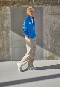 Jonge persoon staat binnen en draagt een felblauwe jas, beige broek en witte sneakers, tegen een textuurgrijze muur met zonlicht.