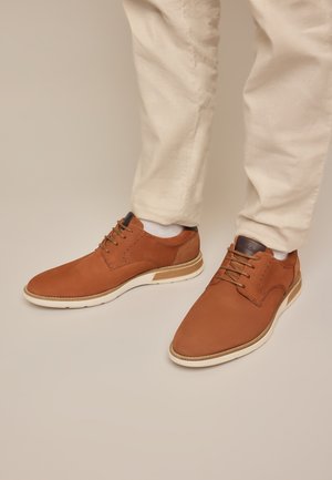 Braune Leder-Schnürschuhe getragen mit weißen Socken und beigen Hosen vor neutralem Hintergrund.