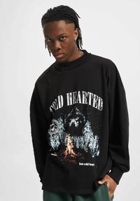 Melna pārsteigtā džemperis ar grafiku, kurā redzams lācis, koki un uguns; virs attēla ir uzraksts "COLD HEARTED" treknrakstā.