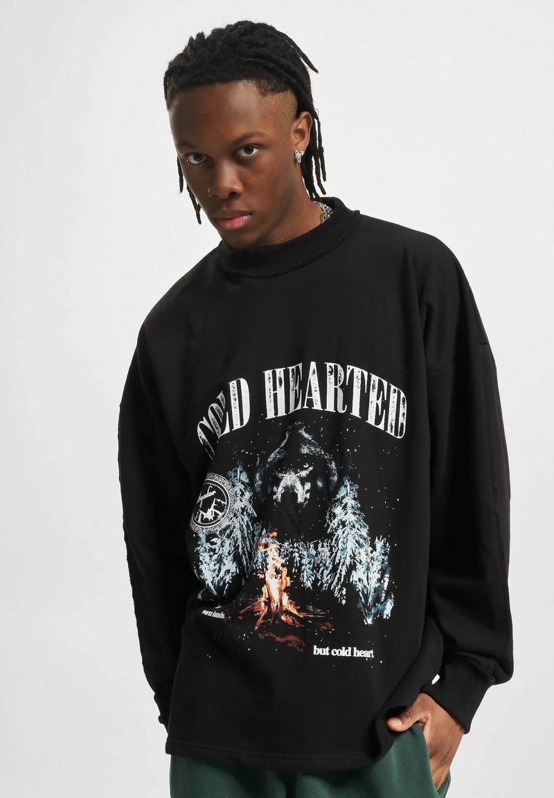 Melna pārsteigtā džemperis ar grafiku, kurā redzams lācis, koki un uguns; virs attēla ir uzraksts "COLD HEARTED" treknrakstā.
