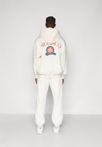 Κρέμα φούτερ με την ένδειξη "MONACO" σε πορτοκαλί στο πίσω μέρος, με κυκλικό λογότυπο. Συνδυάζεται με ασορτί κρέμα φόρμες και λευκά αθλητικά παπούτσια.