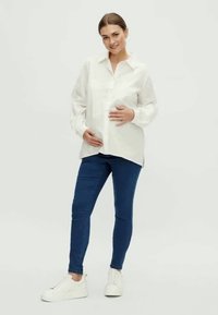 Bela srajca na gumb, modri oprijeti džins in bele superge. Model nežno drži okroglo trebuh, nevtralna ozadje.