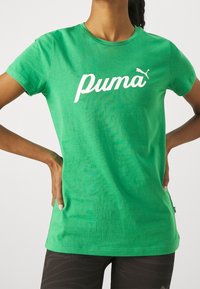 Osoba v jasně zeleném tričku Puma s bílým logem, ruce položené na bocích, v kombinaci s tmavými vzorovanými kalhotami.