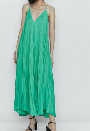 Vrouw draagt een vloeiende, geplooide teal maxi-jurk met dunne bandjes en een diepe V-hals, gecombineerd met zwarte, open sandaal hakken met bandjes.