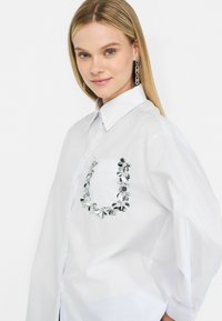 Bílá košile s knoflíky a strukturovaným límcem, v oversized střihu, se dekorativní stříbrnou květinovou aplikací na náprsní kapse.
