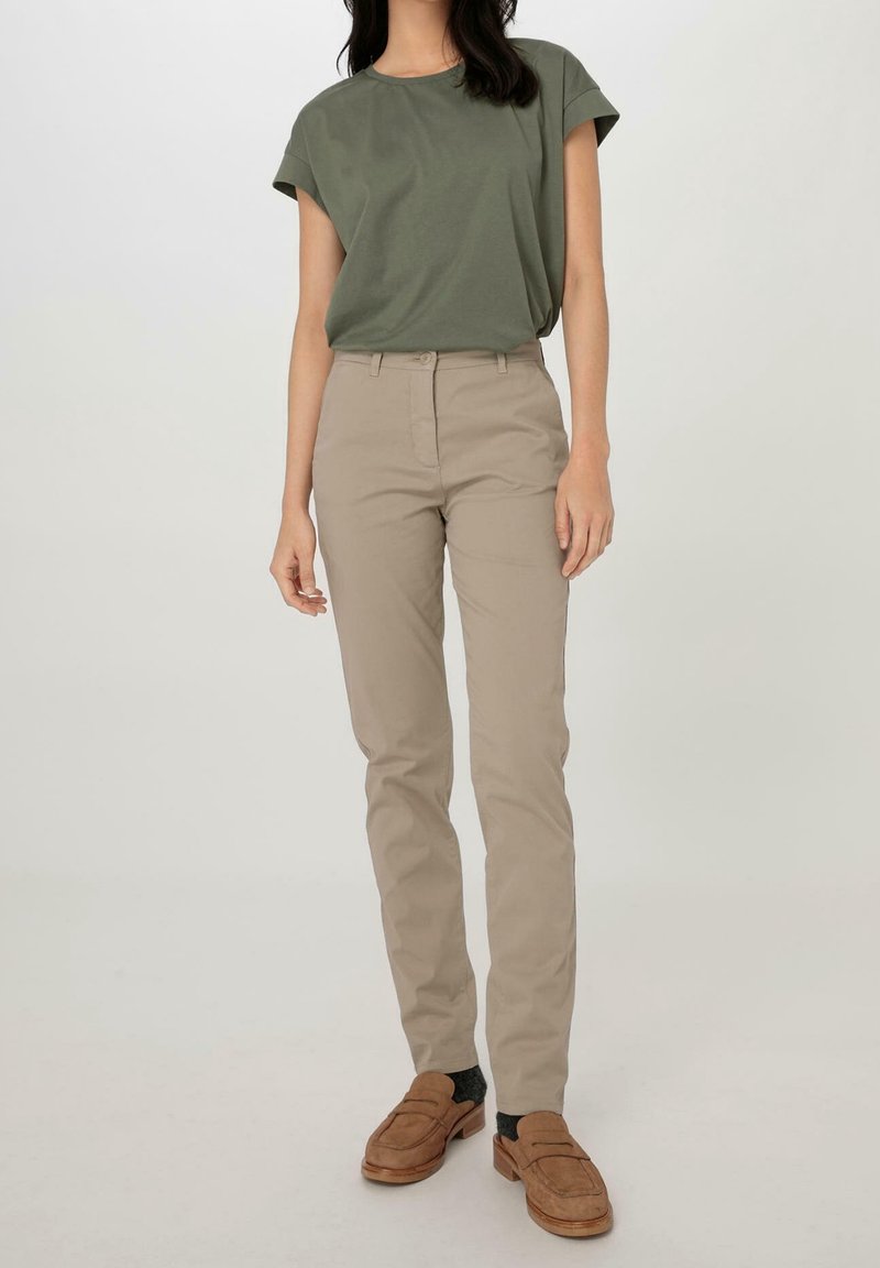 Femme portant un haut manches courtes vert olive, un pantalon slim beige, des chaussettes foncées et des mocassins marron, debout devant un fond clair uni.