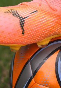 Helle orange Fußballschuhe mit strukturierter Oberfläche, schwarzem Logo und gelben Akzenten, positioniert auf einem orange-grauen Fußball mit Musterdetails.