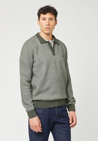 Man in een groen, gestreept, langarm polo-sweater en donkerblauwe jeans, staand tegen een effen lichte achtergrond.