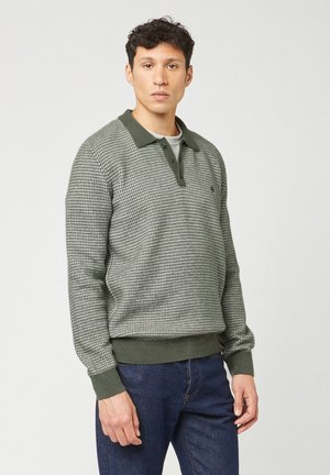 Man in een groen, gestreept, langarm polo-sweater en donkerblauwe jeans, staand tegen een effen lichte achtergrond.