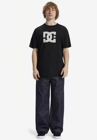 Svart bomulls t-skjorte med en fremtredende hvit DC-logo. Kombinert med løstsittende mørkeblå jeans i denim og svarte joggesko.