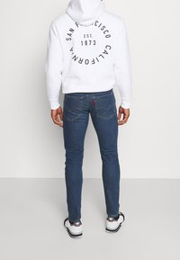 Man som bär en vit hoodie med texten "San Francisco California EST. 1873" på ryggen, blå jeans och vita Adidas-sneakers, står med ryggen vänd.