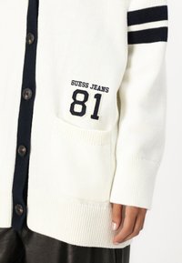 Biały dziergany kardigan z granatowymi dodatkami, z kieszenią ozdobioną haftem "GUESS JEANS" i "81" oraz ciemnymi guzikami. Mankiety i dolna część wykończone ściągaczem.