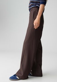 Bruine wijde broek met hoge taille en voorplooien, gemaakt van gladde stof. Gecombineerd met blauw-witte sneakers.