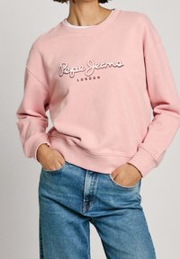 Sudadera rosa con puños y cuello acanalados, con la inscripción "Pepe Jeans London" en letras grandes centrada en el pecho. Combinada con vaqueros azules.