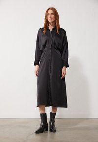 Abito camicia in satin nero con bottoni frontali, maniche lunghe e vestibilità rilassata. Abbinato a stivali neri con tacco alla caviglia. Design semplice.