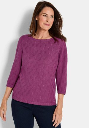 Lila gestrickter Pullover mit rundem Ausschnitt, drei Viertelärmeln, strukturiertem Muster und gerippten Bündchen und Saum. Kombiniert mit dunklen Jeans.