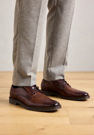 Brown leather dress shoes worn with light gray tailored trousers on a wooden floor.