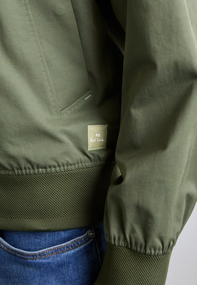 Oliivinvihreä bombere takki, jossa on teksturoitu neulehelma, sivutasku ja etiketti, jossa lukee "PS Paul Smith". Liukas kangaspinta.