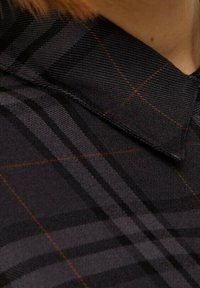 Närbild av en mörk rutig skjortkrage med röda och grå linjer, bärs av en person med ljus hy och ljusbrunt hår.