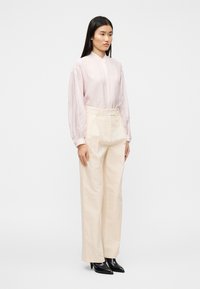 Ανοιχτό ροζ πουκάμισο μακρυμάνικο με ψηλό γιακά, συνδυασμένο με κρεμ παντελόνι φαρδύ και μαύρες μυτερές γόβες· απαλό ύφασμα με διακριτική υφή.