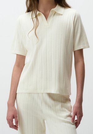Kvinde iført en cremefarvet ribbet kortærmet poloshirt og matchende ribbede bukser mod en ensfarvet baggrund.